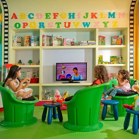 DAMAC Maison Dubai Mall Street Exterior foto The photo shows a colorful children's playroom. In the foreground, three children are sitting in bright green chairs, focused on a television that displays a children's show. The walls are decorated with large, colorful letters from A to Z. Behind th
