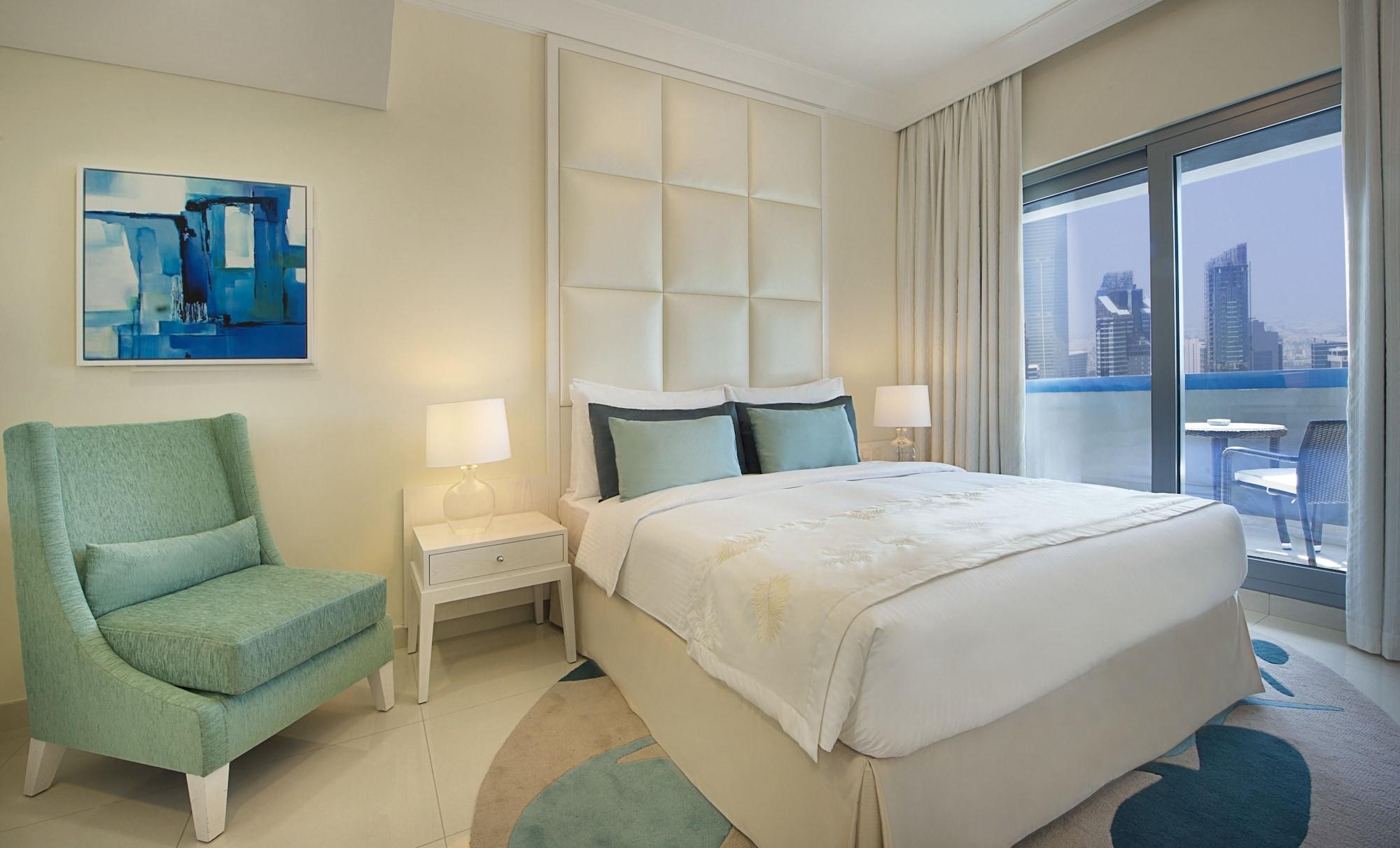 DAMAC Maison Dubai Mall Street Exterior foto The photo shows a modern hotel room with a stylish design. It features a bed with a soft, light-colored comforter and matching decorative pillows. Beside the bed, there are two bedside lamps providing soft lighting. A comfortable green armchair is po