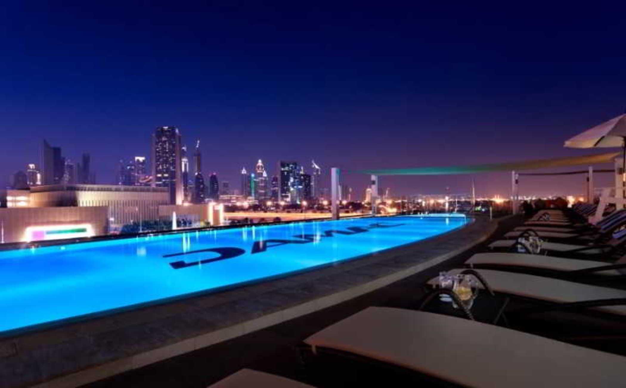 DAMAC Maison Dubai Mall Street Exterior foto The photo depicts a stunning rooftop pool area illuminated at night. The pool has a vibrant blue color and reflects the city lights, creating a serene atmosphere. In the background, the skyline of a modern city is visible, showcasing tall buildings a