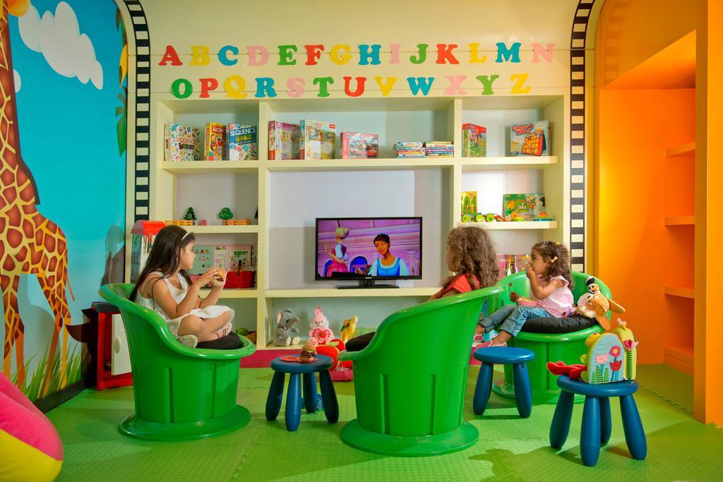 DAMAC Maison Dubai Mall Street Exterior foto The photo shows a colorful children's playroom. In the foreground, three children are sitting in bright green chairs, focused on a television that displays a children's show. The walls are decorated with large, colorful letters from A to Z. Behind th