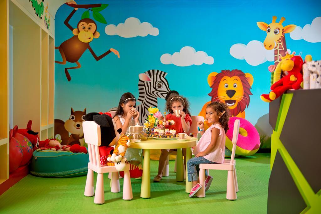 DAMAC Maison Dubai Mall Street Exterior foto The photo shows a colorful playroom where three children are seated at a small round table. They appear to be enjoying snacks or refreshments. The background features vibrant, cartoon-style decorations, including a monkey, a zebra, and a lion, all of