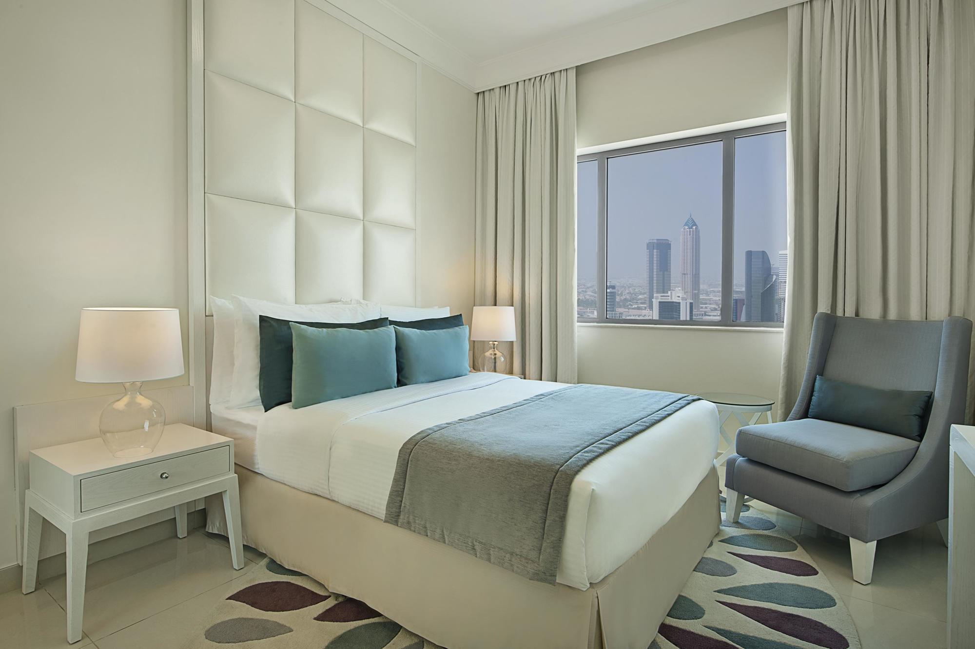 DAMAC Maison Dubai Mall Street Exterior foto The photo shows a modern hotel room. It features a neatly made bed with white linens and two decorative pillows in shades of blue. A light-colored headboard is positioned behind the bed, and there are floor-to-ceiling curtains that let in natural lig