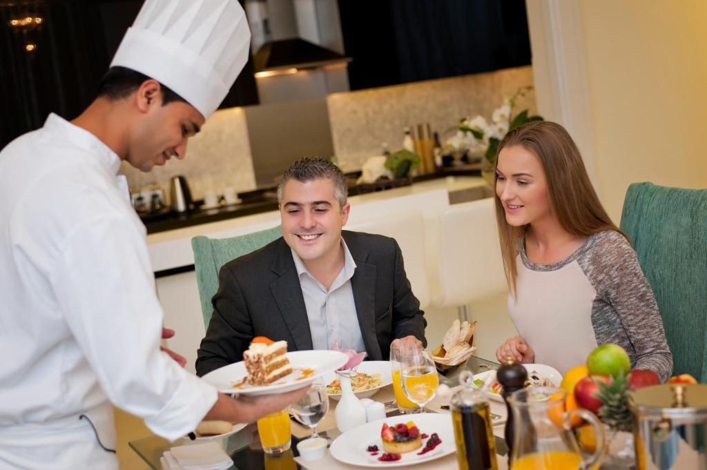 DAMAC Maison Dubai Mall Street Exterior foto The photo shows a dining scene where a chef, wearing a white chef's hat and attire, is serving food to a couple seated at a table. The man, dressed in a suit, has a smile on his face as he receives a dessert plate, while the woman, wearing a casual t