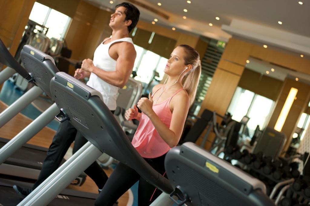 DAMAC Maison Dubai Mall Street Exterior foto The photo shows two individuals jogging on treadmills in a gym. The man, wearing a white tank top, appears focused as he runs. The woman, dressed in a pink workout top, is also jogging beside him. The gym environment has modern equipment, with a well