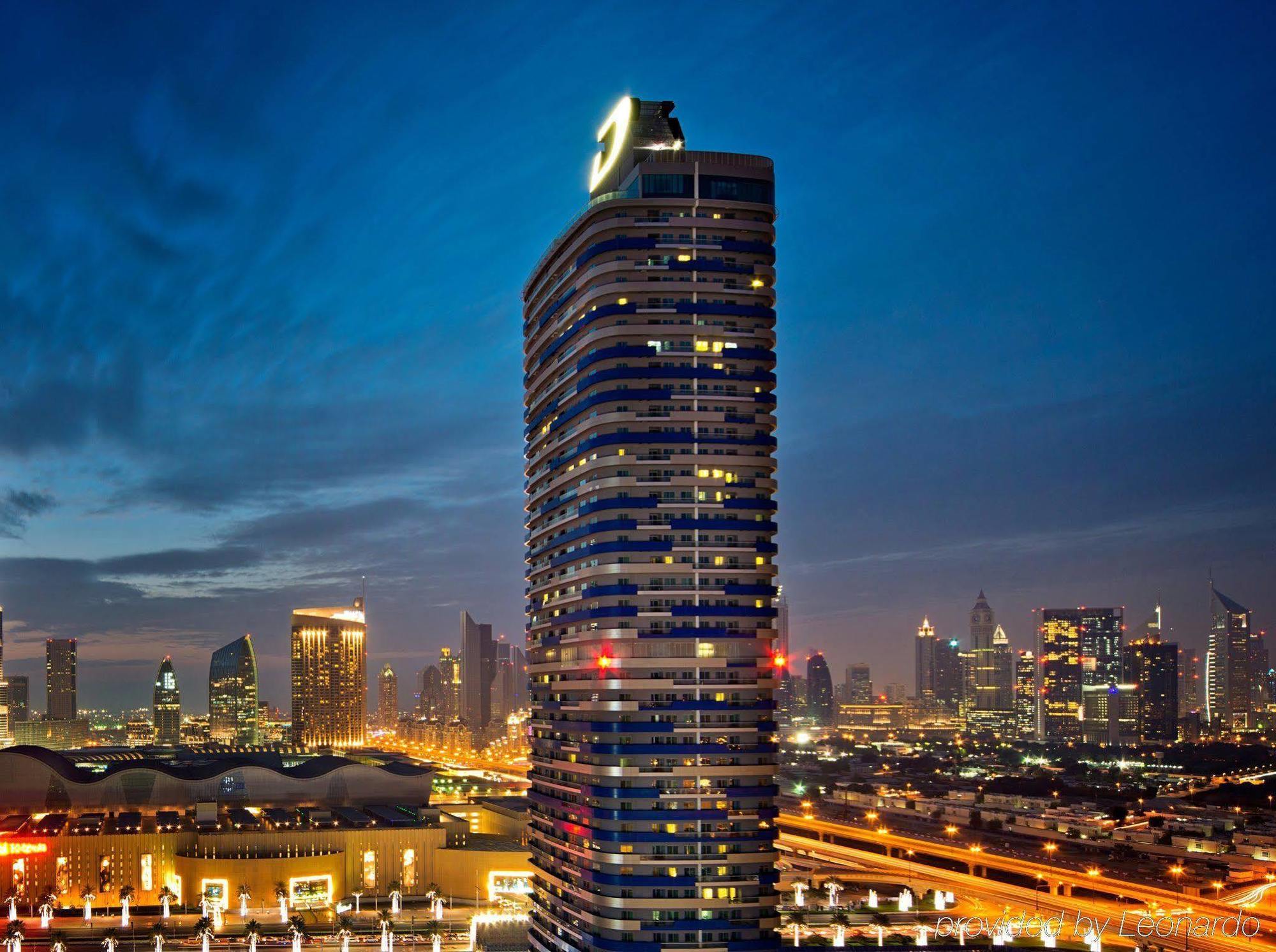 DAMAC Maison Dubai Mall Street Exterior foto The photo shows a tall, modern skyscraper illuminated against a twilight sky. The building features a sleek, curved design with multiple floors and a prominent logo or symbol at the top. In the background, the city skyline is visible, showcasing a mi