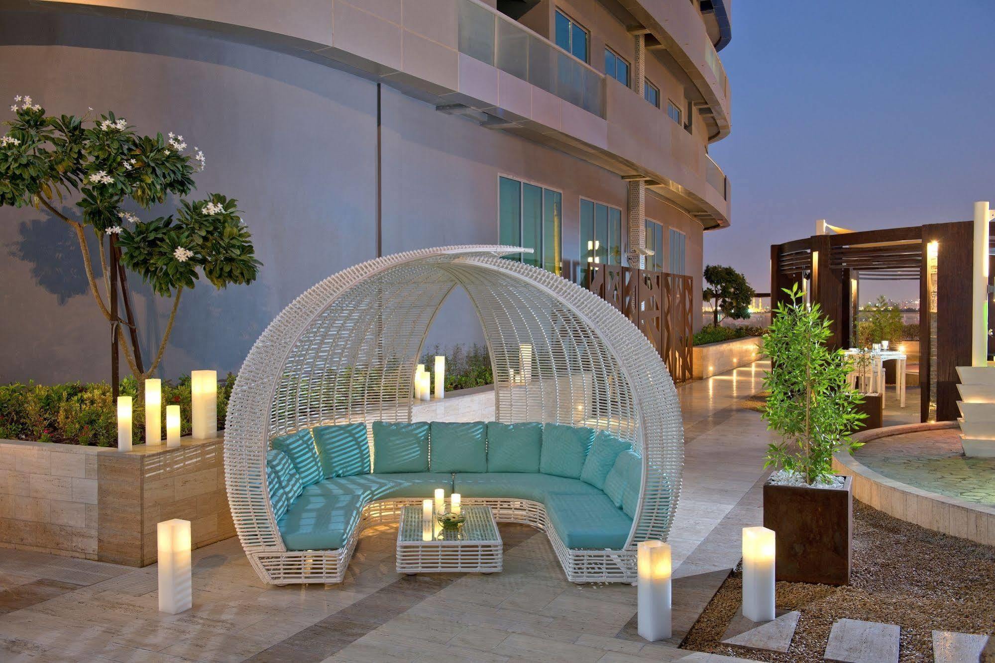 DAMAC Maison Dubai Mall Street Exterior foto The photo shows a modern outdoor seating area featuring a unique, rounded structure made of white wicker. Inside, there is a spacious, cushioned seating arrangement with light blue cushions. The setting is well-lit, with several tall, elegant candles
